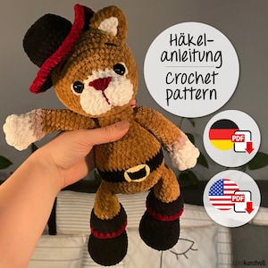 Puede incluir: Patrón de crochet para un oso de peluche marrón que lleva un sombrero rojo y negro y un cinturón negro. El patrón está disponible en inglés y alemán.