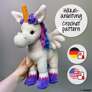 May include: A white crocheted unicorn plush toy with a yellow horn, purple and blue mane, and white wings. The feet are accented with purple and pink yarn. Text on the image reads Häkelanleitung crochet pattern with German and American flag icons.