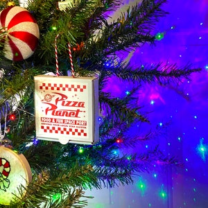 May include: A red and white striped candy cane ornament hangs on a Christmas tree with a Pizza Planet box ornament. The box is red and white checkered with the words "Pizza Planet" in a white circle with a red star. The text below reads "Food & Fun Space Port Serving Your Local Star Cluster".