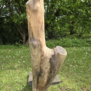 May include: A wooden sculpture, resembling a tree trunk, stands on a square base in a grassy area. The sculpture is light brown with darker streaks and a smooth, angled top. The side branch is cut flat. The sculpture is outdoors.