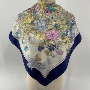 May include: A square silk scarf featuring a floral print with shades of pink, yellow, blue, and green on a cream background. The scarf has a wide navy blue border with a thin pink inner stripe and the Gucci brand name printed in the lower corner.