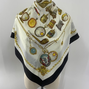 May include: A square silk scarf featuring a white background with a black border and a gold chain pattern. The print includes various vintage pocket watches, a wooden pipe, and decorative canisters. The text La Fenice is printed in the corner.