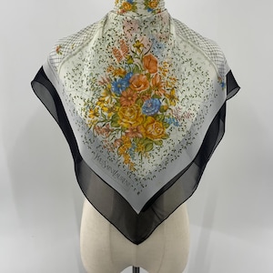 May include: Square silk scarf with a floral design in shades of yellow, orange, and blue. The scarf has a white background with a black border and the text "YVES SAINT LAURENT".
