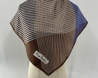 Puede incluir: Un pañuelo cuadrado de seda con un patrón geométrico de rayas y cuadrículas en marrón, blanco y azul. El pañuelo tiene un borde marrón y una etiqueta rectangular blanca en la esquina con el texto Pierre Cardin impreso en color negro.
