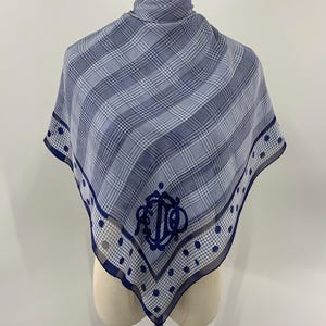 Może przedstawiać: Niebiesko-biała szal z kratą z granatową obwódką i powtarzającym się wzorem w kropki. Szal posiada granatowy monogram pośrodku. Szal jest prezentowany na manekinie.