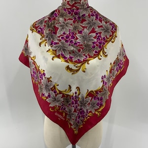 Puede incluir: Un pañuelo cuadrado Givenchy con borde rojo oscuro y centro color crema. El diseño muestra racimos de uvas moradas y hojas grises, acentuados por patrones de volutas doradas. La palabra GIVENCHY está impresa en una esquina del pañuelo.