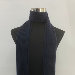 Peut inclure: Une écharpe en tricot côtelé bleu foncé avec une bordure marron, grise et bleue. L'écharpe porte une petite étiquette blanche avec le texte "Woolrich" dessus.