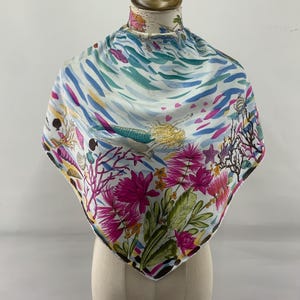 Könnte beinhalten: Ein quadratischer Schal mit einem Meerjungfrau- und Blumendesign. Der Schal hat einen weißen Hintergrund mit einer farbenfrohen Illustration einer Meerjungfrau, rosa Blumen und Korallen. Die Ränder sind schwarz.
