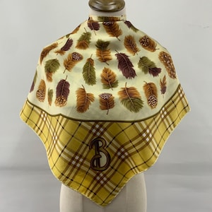 May include: Square scarf with a feather pattern in shades of brown, green, and gold on a cream background. The border features a yellow and brown plaid design. A brown monogram "B" is embroidered on the scarf.