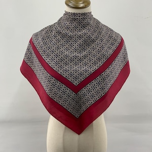 Puede incluir: Un pañuelo cuadrado con un patrón geométrico en azul marino, beige y rojo. El pañuelo se exhibe en un maniquí, mostrando su diseño y forma. El borde es de color rojo liso, enmarcando el centro estampado.