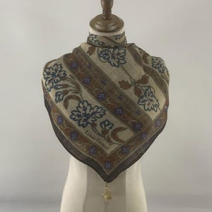 以下が含まれることがあります： ベージュ、ブラウン、ブルーの花柄の正方形のスカーフ。 スカーフは「Louis Feraud」の文字と金色のチャームが付いたストライプのデザインです。 スカーフはマネキンに展示されています。