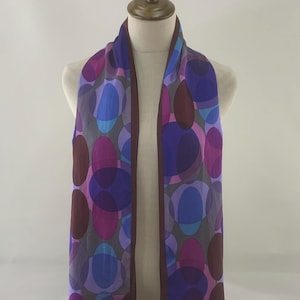 Puede incluir: Una bufanda larga y rectangular con un patrón de óvalos superpuestos en tonos morados, azules y granates. La bufanda se exhibe en un maniquí y tiene un borde morado oscuro. El fondo es blanco liso.