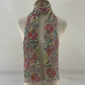 Puede incluir: Bufanda floral con fondo crema, adornada con flores rojas, rosas, amarillas y moradas, y hojas verdes. La bufanda está drapeada sobre un maniquí, mostrando su forma rectangular y bordes deshilachados. Un accesorio elegante.