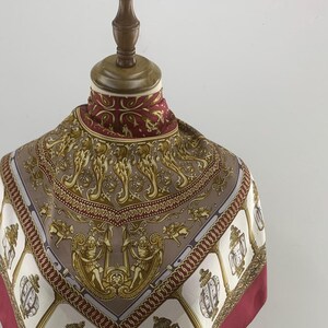 May include: A red, white and gold silk scarf with a patterned design featuring ornate details, cherubs, and a repeating motif of lanterns. The scarf is displayed on a mannequin and has a label that reads "Louis Quintiny".