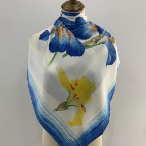 May include: Square white scarf with a blue and yellow floral design. The scarf features large blue iris flowers and a yellow iris flower. A blue border frames the scarf.