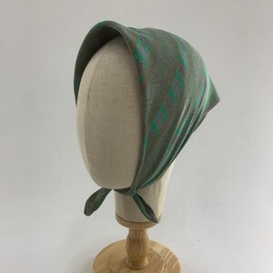 Puede incluir: Un pañuelo con estampado de rayas verdes y grises, atado al cuello, expuesto sobre una cabeza de maniquí. El pañuelo presenta rayas verticales en tonos verdes, grises y marrones. La cabeza del maniquí es beige y está sobre un soporte de madera.