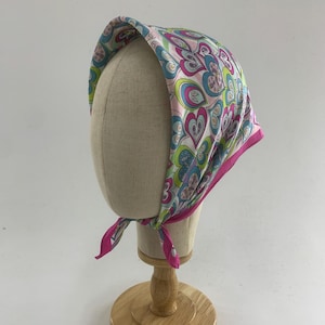 Puede incluir: Un pañuelo colorido con un diseño de corazones en rosa, azul y verde, con un borde rosa. El pañuelo está atado al cuello y se muestra en una cabeza de maniquí. El diseño del pañuelo es llamativo y vibrante.