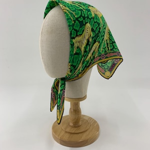 May include: A green silk scarf with a zebra print, displayed on a mannequin head. The scarf features a repeating pattern of yellow zebras against a green background with black spots. The edges are trimmed with a decorative border.