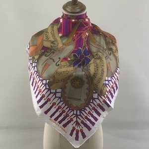 Puede incluir: Un pañuelo cuadrado de seda con un diseño de instrumentos musicales en tonos beige, naranja, morado y gris. El pañuelo tiene un borde blanco con un patrón repetido de borlas moradas. El texto "ROCHAS-PARIS" está impreso en el borde.