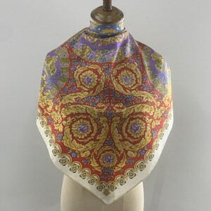 May include: A square silk scarf with a vibrant baroque design. The scarf features a red central field with gold floral and scroll motifs, bordered by a cream-colored edge. Purple and blue floral accents add color to the design.