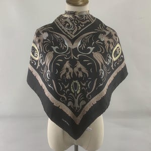 Puede incluir: Pañuelo cuadrado negro con un diseño ornamentado con pájaros, elementos florales y volutas en tonos beige y plateado. El pañuelo se exhibe en un maniquí y tiene un borde con flecos.