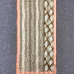 Op de afbeelding: Rechthoekige sjaal met een patroon. De sjaal heeft verticale strepen in tinten beige en bruin, met een ruitpatroon aan de rechterkant. De randen zijn afgewerkt met een perzikkleurige rand. De sjaal is gemaakt van een lichtgewicht materiaal.
