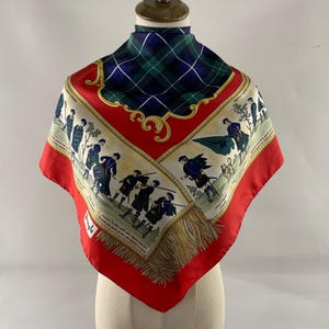 May include: A square silk scarf with a red border and a green and blue tartan pattern. The scarf features illustrations of Scottish soldiers in kilts and a gold fringe. The brand name "Pringle" is visible.