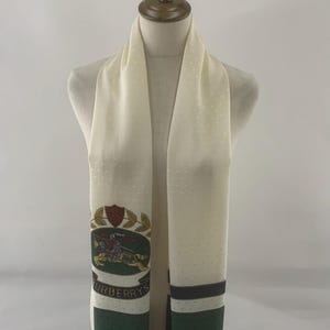 May include: Cream-colored scarf with a subtle polka dot pattern. Features a green and white logo with the word "BURBERRYS". The scarf has a green dotted border and a dark stripe near the bottom. A classic accessory.