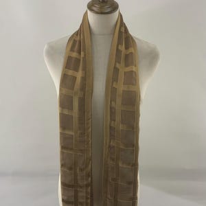 Puede incluir: Una bufanda color beige con un diseño de rejilla, exhibida en un maniquí. La bufanda tiene un diseño rectangular con cuadrados y líneas de color beige más oscuros. La bufanda está hecha de un material suave y fluido.