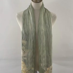 May include: A light green scarf with a floral design. The scarf is draped over a mannequin. The floral design features yellow roses and green leaves. The scarf has a vertical striped pattern.