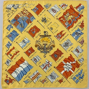 May include: A yellow silk scarf featuring a repeating pattern of flags, each with a unique design and color scheme. The flags are arranged diagonally across the scarf, with text labels. A central emblem includes an anchor and the word "PAVOIS".