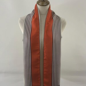 Puede incluir: Bufanda de seda con un estampado de rayas verticales en negro, blanco y naranja. La bufanda se exhibe en un maniquí. La bufanda tiene un panel central naranja liso y un borde negro en la parte inferior. La marca "Echo" es visible.