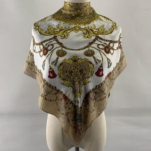 Puede incluir: Un pañuelo de seda con un diseño decorativo. El pañuelo presenta un fondo blanco con detalles dorados, marrones y rojos. El diseño incluye patrones ornamentados, borlas y detalles de cadenas. El pañuelo se exhibe en un maniquí.