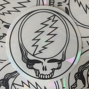 May include: A white, iridescent sticker with a black outline of a skull with a lightning bolt in the middle. The skull is smiling and has a wavy, flowing hair design.