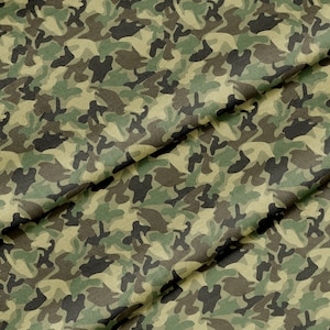 May include: A close-up of a camouflage fabric with a green, brown, and beige pattern. The fabric is folded in half, creating a diagonal line across the image.
