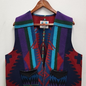 May include: A sleeveless vest featuring a vibrant geometric pattern in shades of red, navy blue, purple, and teal. The garment includes two front pockets with black leather-like trim and a label at the inner neckline that reads Wooded River.