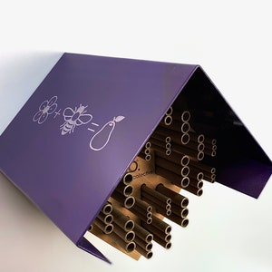 May include: Purple metal bee house with a brown cardboard insert containing multiple round holes for bees to nest in. The house has a white graphic of a flower, bee, and eggplant with the text "o. collective".