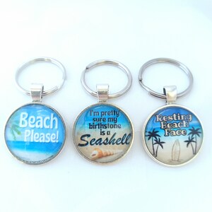 May include: Three round keychains with different beach-themed designs. The first keychain says "Beach Please!" with a palm tree and ocean scene. The second keychain says "I'm pretty sure my birthstone is a Seashell" with a seashell and ocean scene. The third keychain says "Resting Beach Face" with palm trees and a surfboard.