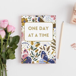 May include: A white notebook with a floral pattern and butterflies. The notebook has the text "ONE DAY AT A TIME" in gold lettering.