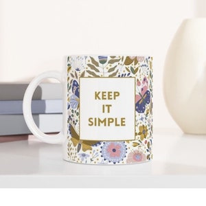 May include: White ceramic mug with a floral pattern in blue, pink, and gold. The mug features the text "KEEP IT SIMPLE" in a gold frame. The mug is on a white surface with books and vases in the background.