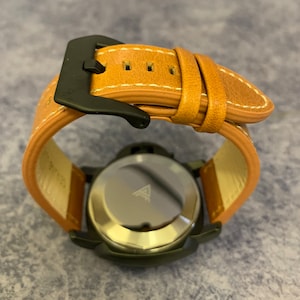 K&ouml;nnte beinhalten: Eine schwarze Uhr mit einem braunen Lederarmband. Die Uhr hat ein silbernes Zifferblatt mit einem Dreieckslogo. Die Uhr liegt auf einer braunen Oberfl&auml;che.