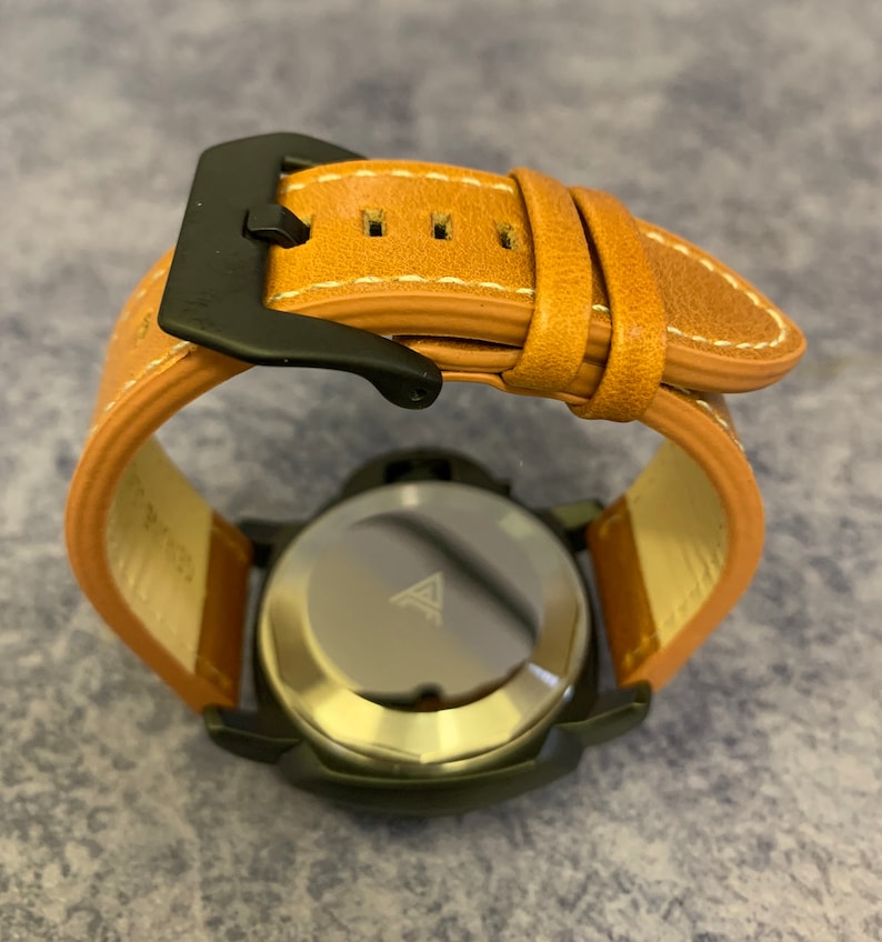 K&ouml;nnte beinhalten: Eine schwarze Uhr mit einem braunen Lederarmband. Die Uhr hat ein silbernes Zifferblatt mit einem Dreieckslogo. Die Uhr liegt auf einer braunen Oberfl&auml;che.