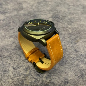 K&ouml;nnte beinhalten: Eine schwarze Uhr mit einem braunen Lederarmband. Das Zifferblatt der Uhr hat einen schwarzen Hintergrund mit wei&szlig;en Zahlen und einem goldenen Zeiger. Auf dem Zifferblatt der Uhr steht auch das Wort "ARCHIVE".