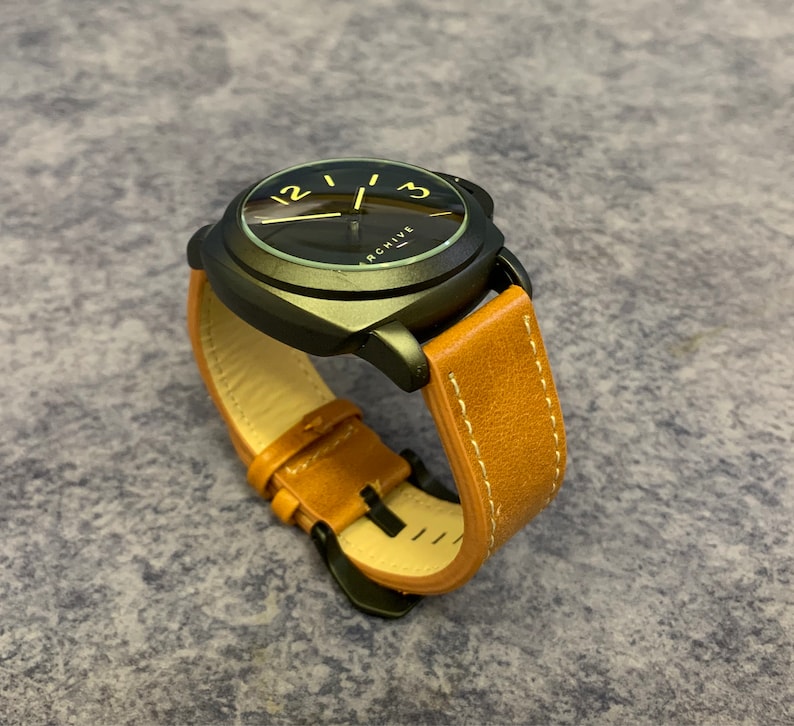 K&ouml;nnte beinhalten: Eine schwarze Uhr mit einem braunen Lederarmband. Das Zifferblatt der Uhr hat einen schwarzen Hintergrund mit wei&szlig;en Zahlen und einem goldenen Zeiger. Auf dem Zifferblatt der Uhr steht auch das Wort "ARCHIVE".