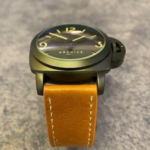 K&ouml;nnte beinhalten: Eine schwarze Armbanduhr mit einem braunen Lederarmband. Das Zifferblatt der Uhr hat einen schwarzen Hintergrund mit gelben Zahlen und Zeigern. Auf dem Zifferblatt der Uhr steht auch das Wort "ARCHIVE".