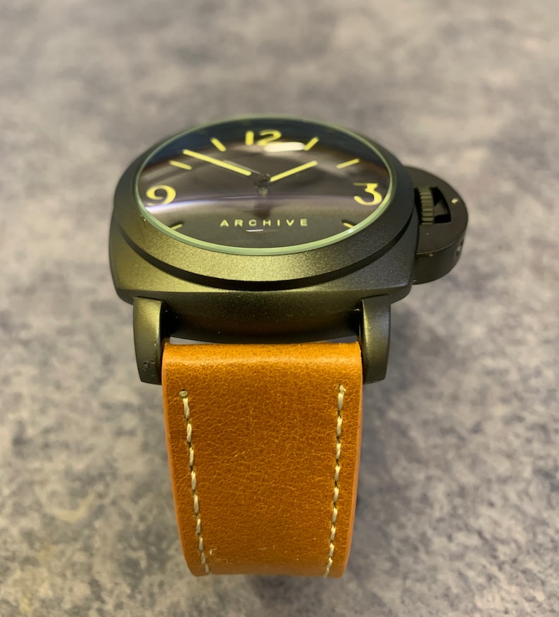 K&ouml;nnte beinhalten: Eine schwarze Armbanduhr mit einem braunen Lederarmband. Das Zifferblatt der Uhr hat einen schwarzen Hintergrund mit gelben Zahlen und Zeigern. Auf dem Zifferblatt der Uhr steht auch das Wort "ARCHIVE".
