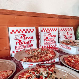 Puede incluir: Varias pizzas con ingredientes como pepperoni, aceitunas y cebollas se muestran en bandejas de metal. Se ven cajas de pizza con el logotipo "Pizza Planet Delivery Shuttle". Las pizzas están sobre una superficie de baldosas rojas.