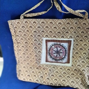 May include: A brown and beige tote bag with a brown and white patterned design. The bag has a square patch with a brown and white design in the center. The bag has two long brown straps.