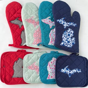 May include: Set of six quilted oven mitts and pot holders featuring a Michigan state outline in various fabric patterns. The patterns include floral, gingham, and blue and white floral. The mitts and holders are in red, teal, green, and navy blue.
