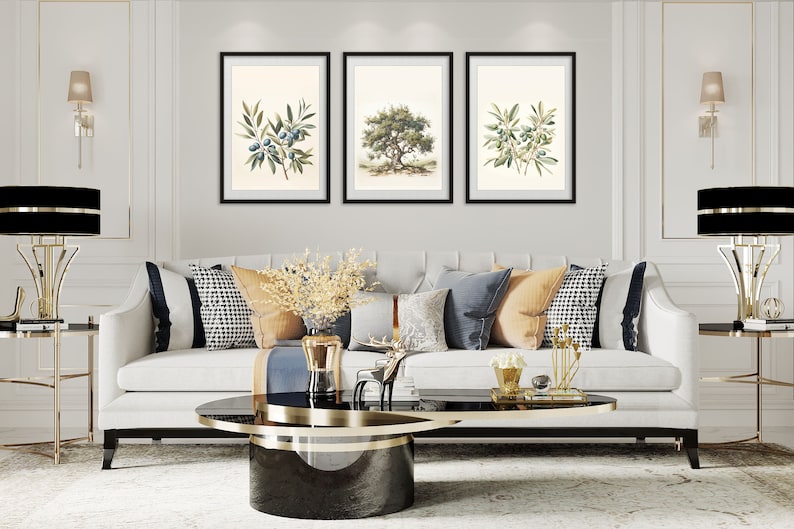 May include: Three framed botanical prints of olive branches and a tree hang above a white sofa with decorative pillows. The room features gold accents, including lamps and side tables, and a black and gold coffee table. The overall style is elegant and classic.